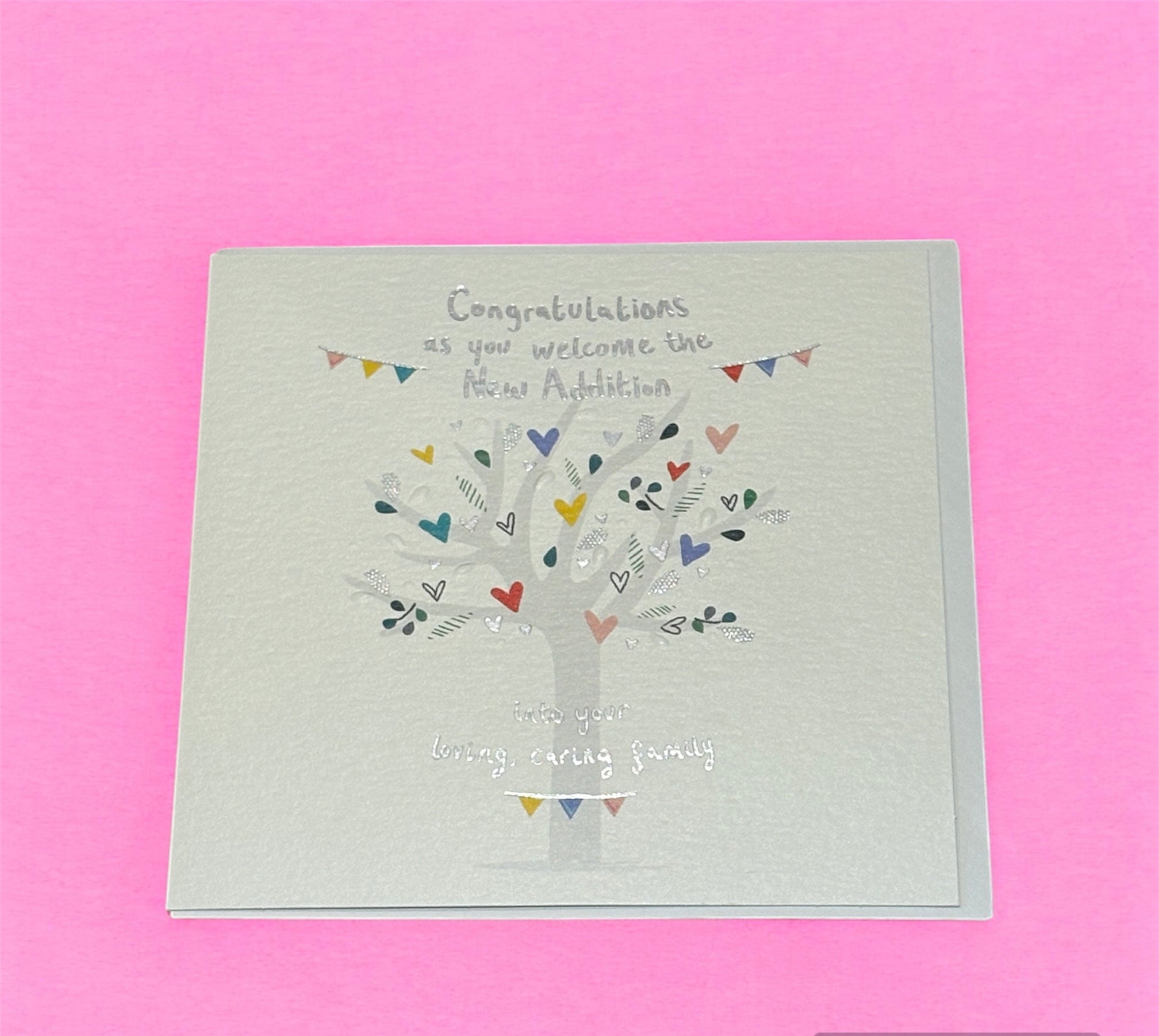 A greeting card with a white background featuring a grey tree with colorful hearts and a string of triangular bunting with the text 'Congratulations as you welcome the New Addition'.