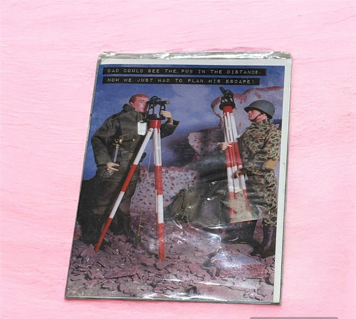 A greeting card with an image of two military figures, one looking through a theodolite mounted on a tripod, and the other carrying a similar tripod. The setting is a destroyed urban landscape with rubble and debris.